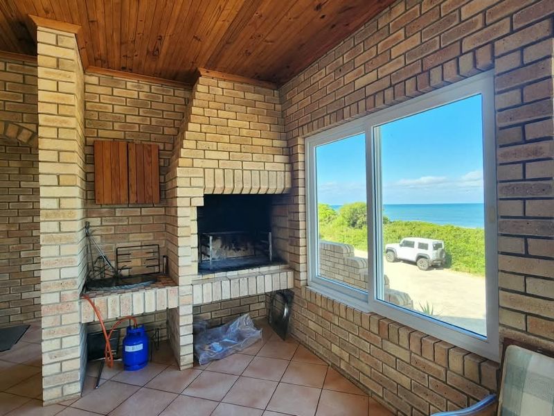 Covered patio featuring a built-in brick braai, timber ceiling, and large windows offering scenic coastal views.