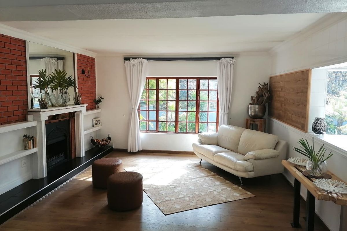 A bright living room featuring timber flooring, a classic fireplace with a brick feature wall, and large timber-framed windows allowing for natural light.