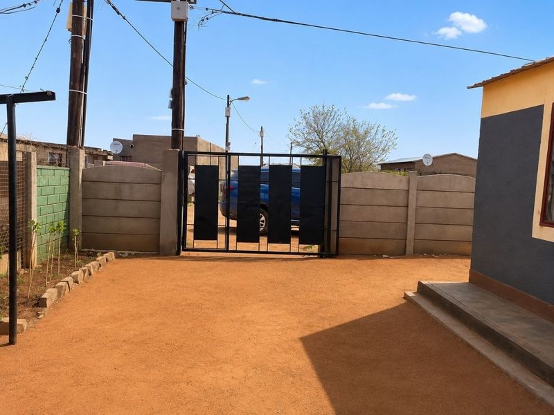Front exterior view showing a metal driveway gate, precast concrete boundary wall, and an unpaved driveway area leading to the house entrance.