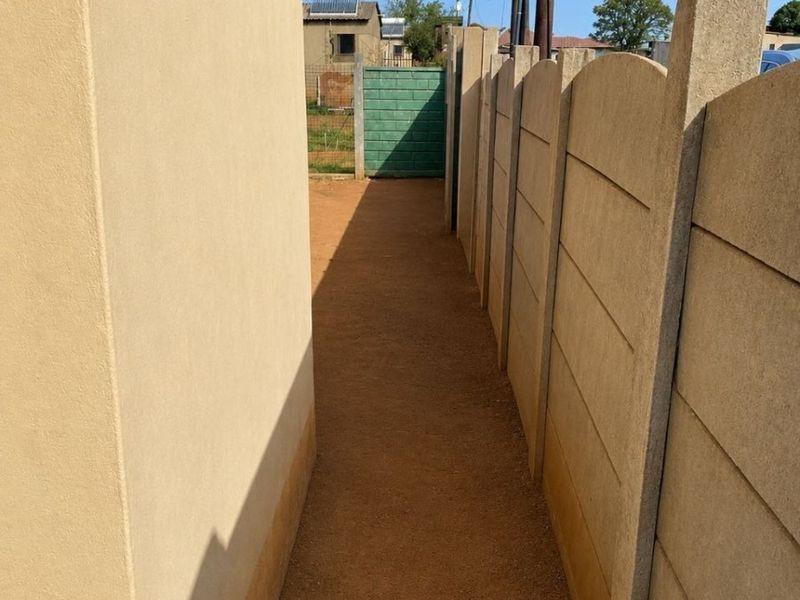 A narrow side passage between a plastered exterior wall and a precast concrete boundary wall with a dirt pathway.
