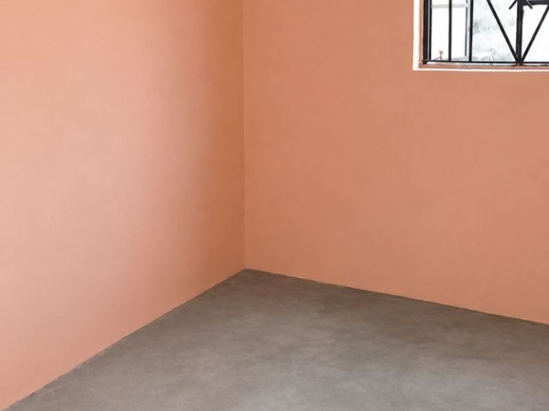 An empty bedroom featuring smooth concrete flooring, peach-toned walls, and a window fitted with security bars.