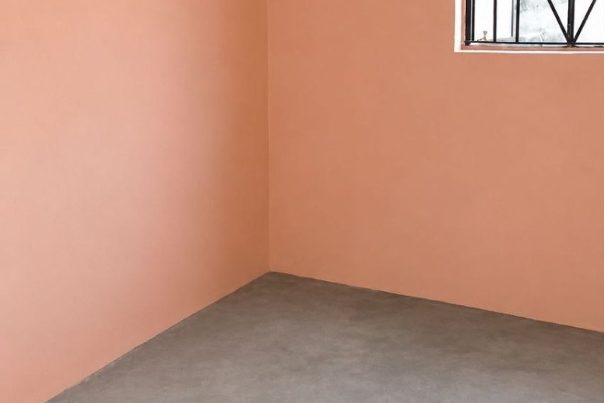 An empty bedroom featuring smooth concrete flooring, peach-toned walls, and a window fitted with security bars.