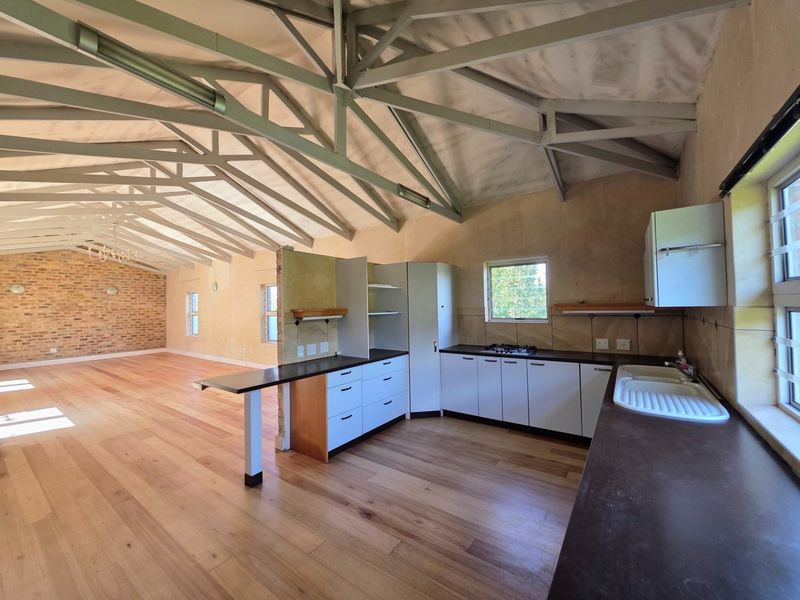 Spacious open-plan living area featuring exposed steel roof trusses, timber flooring, an exposed brick feature wall, and a functional kitchen with built-in cabinetry.