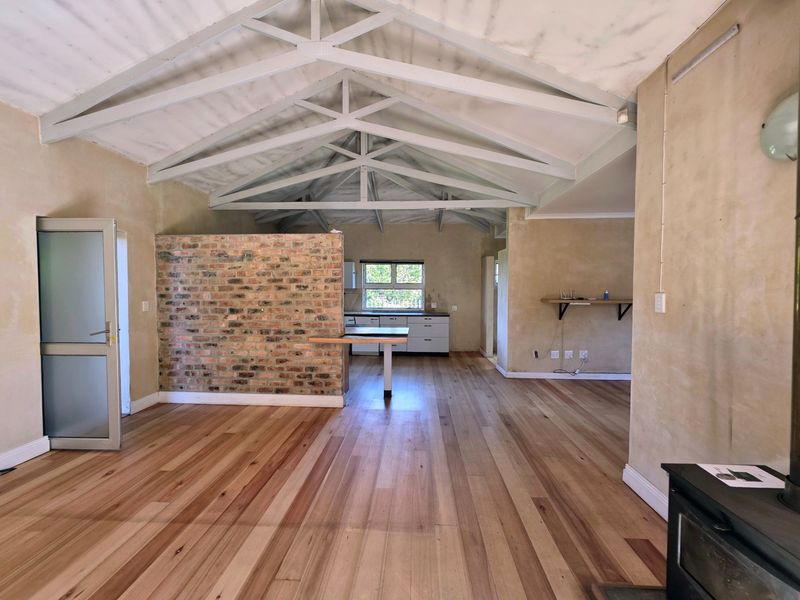 Spacious open-plan living area featuring exposed timber roof trusses, polished timber flooring, and a rustic brick feature wall with an integrated kitchen.
