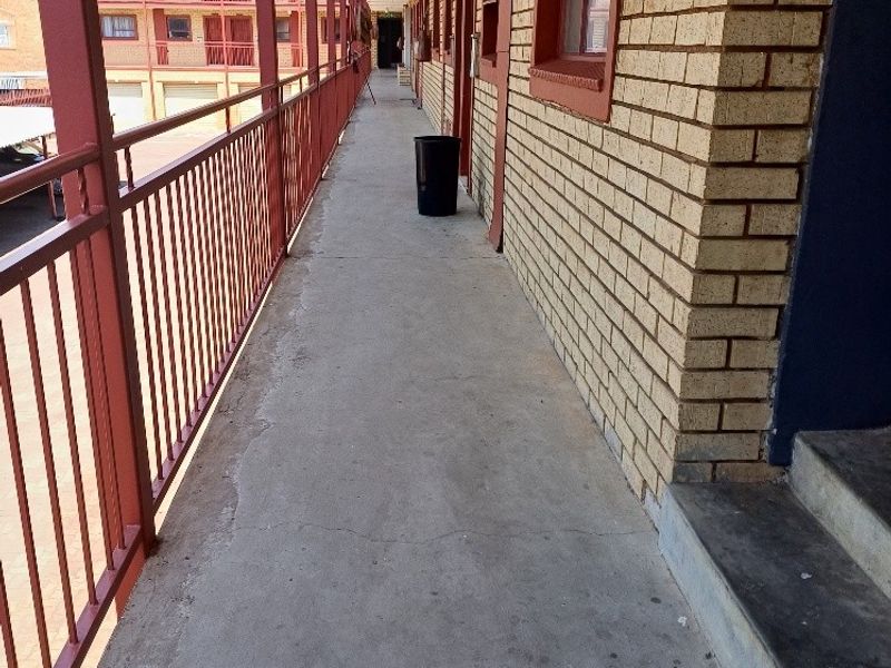 An exterior apartment complex walkway featuring brick walls, metal safety railings, and concrete flooring, providing access to multiple residential units.