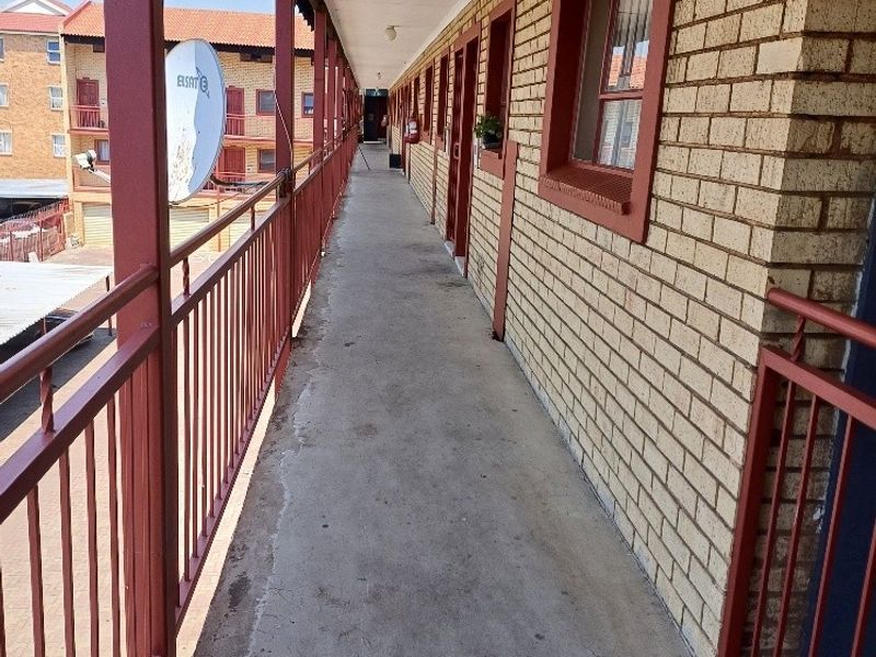 An exterior apartment building walkway featuring brick walls, metal railings, and concrete flooring providing access to multiple residential units.
