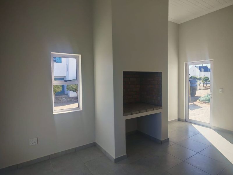 An empty living area featuring a built-in brick braai, neutral tiled flooring, and direct access to the exterior through a glass door.