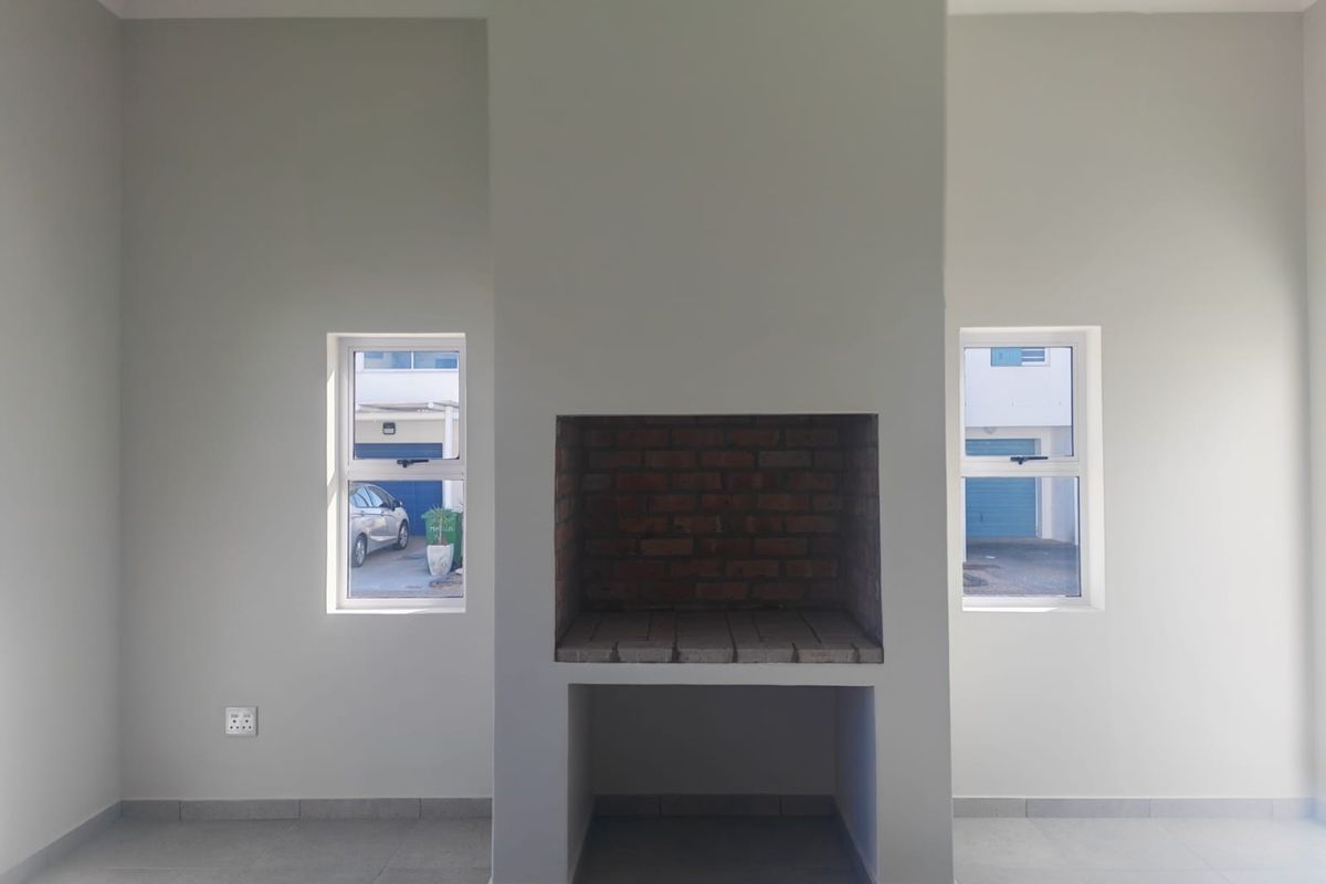 An empty living area featuring a central built-in brick fireplace, neutral tiled flooring, and dual windows providing natural light.