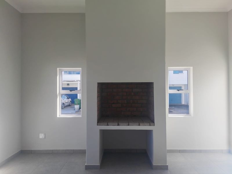 An empty living area featuring a central built-in brick fireplace, neutral tiled flooring, and dual windows providing natural light.