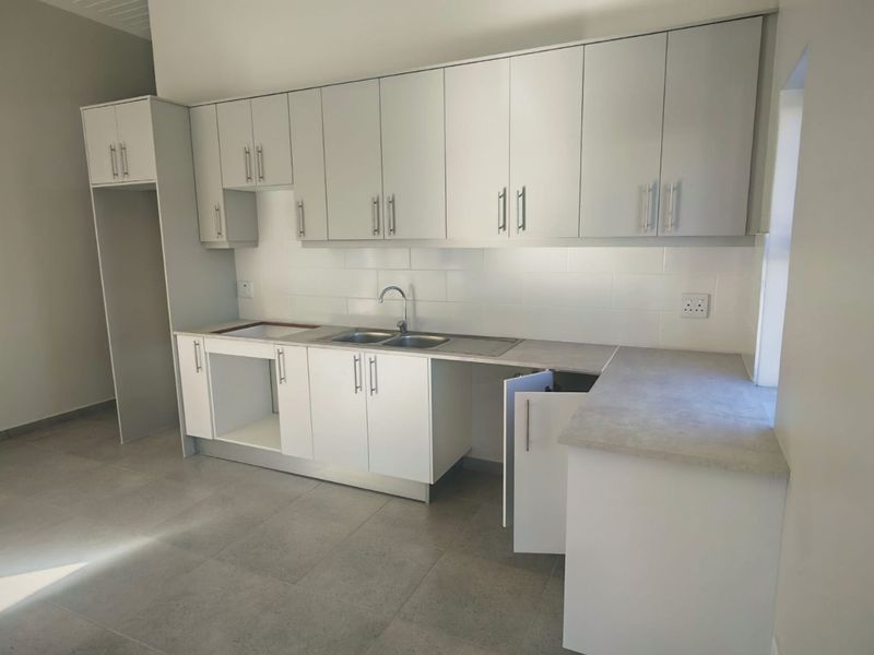 A clean, modern kitchen featuring white built-in cabinetry, grey laminate countertops, a stainless steel sink, and neutral tiled flooring.
