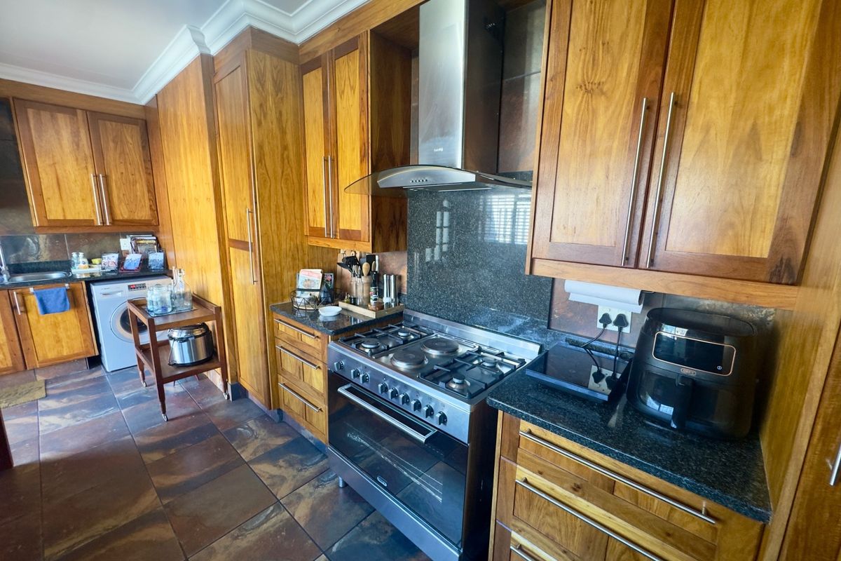A functional kitchen featuring warm-toned wooden cabinetry, granite countertops, stone tile flooring, and a stainless steel gas stove with an integrated extractor fan.