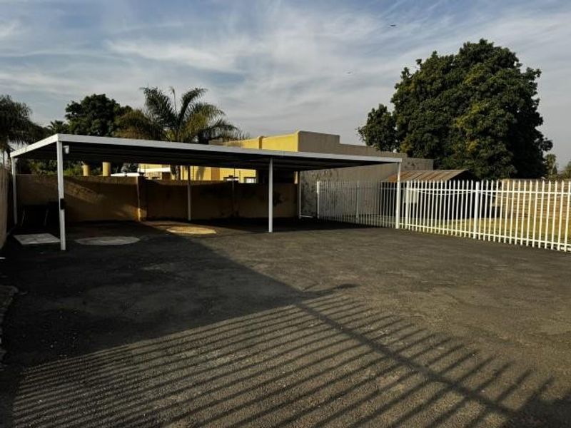 A spacious outdoor parking area featuring a covered carport structure, asphalt driveway, and secure perimeter fencing.