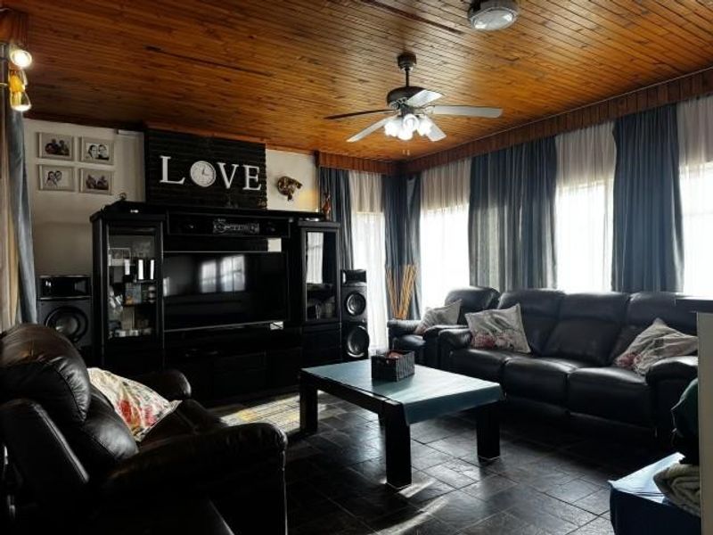 A spacious living room featuring a warm timber tongue and groove ceiling, ceiling fan, tiled flooring, and integrated media cabinetry.
