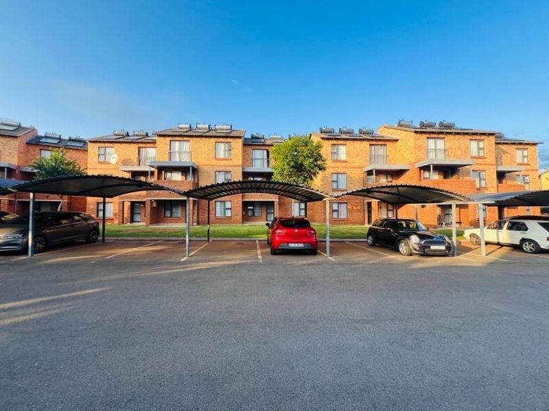 Exterior view of a multi-story face-brick apartment complex featuring covered parking bays and solar water heating installations on the roof.