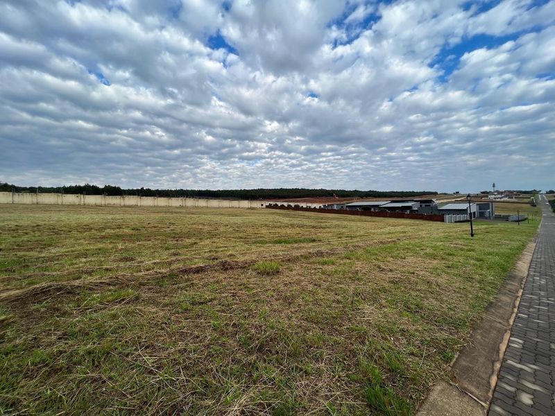 A wide-angle view of a vacant residential plot featuring grass coverage, a perimeter wall, and adjacent paved street access.