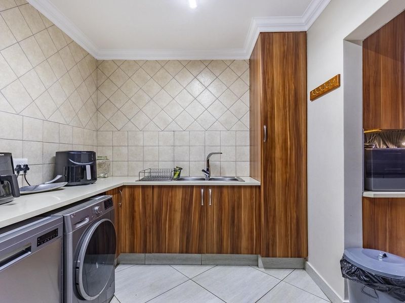 A functional laundry room featuring wood-finish cabinetry, tiled splashbacks, a stainless steel sink, and tiled flooring with space for under-counter appliances.