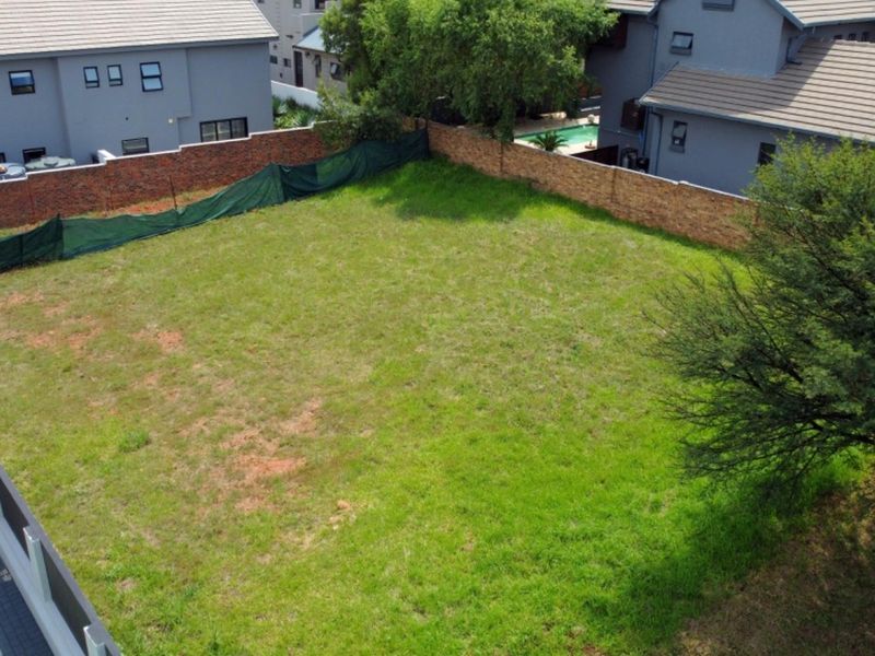 An aerial view of a vacant residential plot featuring a grass lawn, mature tree, and brick boundary walls in a developed neighborhood.