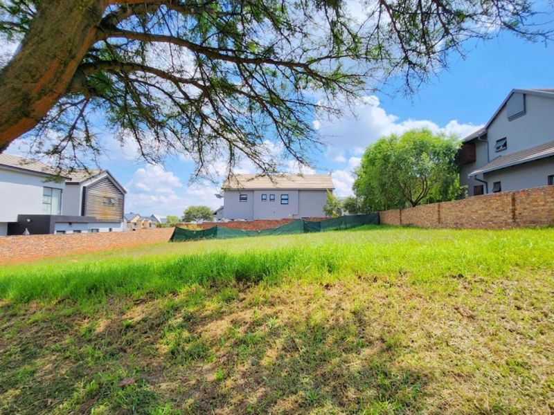 A vacant residential plot featuring a sloped grass surface, enclosed by brick boundary walls and temporary green shade cloth fencing.