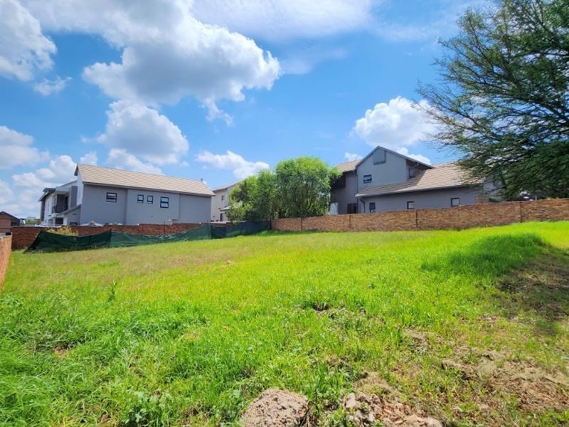 A vacant residential plot featuring a brick boundary wall and green mesh fencing, situated in a developed neighborhood.