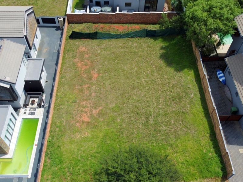 An aerial view of a vacant residential plot situated between existing developed properties, featuring established boundary walls and grass coverage.