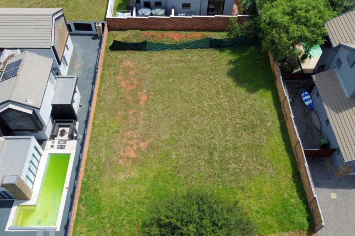 An aerial view of a vacant residential plot situated between existing developed properties, featuring established boundary walls and grass coverage.