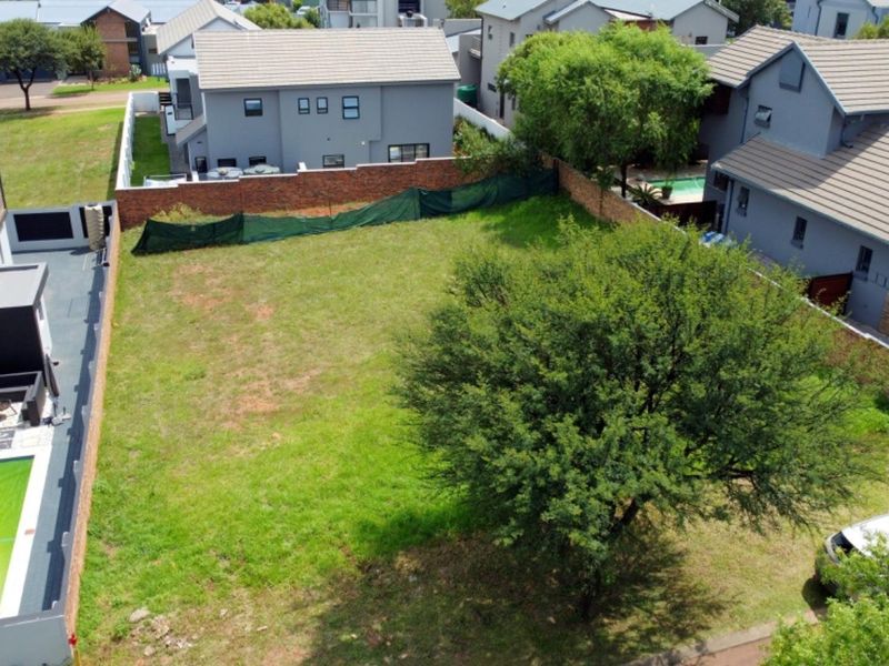 Aerial view of a vacant residential plot featuring a grass lawn, mature tree, and established boundary walls within a developed estate.