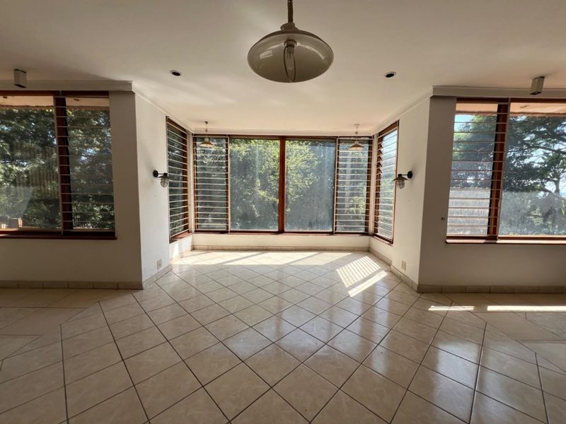 A bright, empty living area featuring large louvered windows, tiled flooring, and a combination of recessed, pendant, and wall-mounted lighting fixtures.