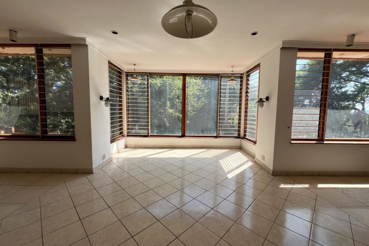 A bright, empty living area featuring large louvered windows, tiled flooring, and a combination of recessed, pendant, and wall-mounted lighting fixtures.