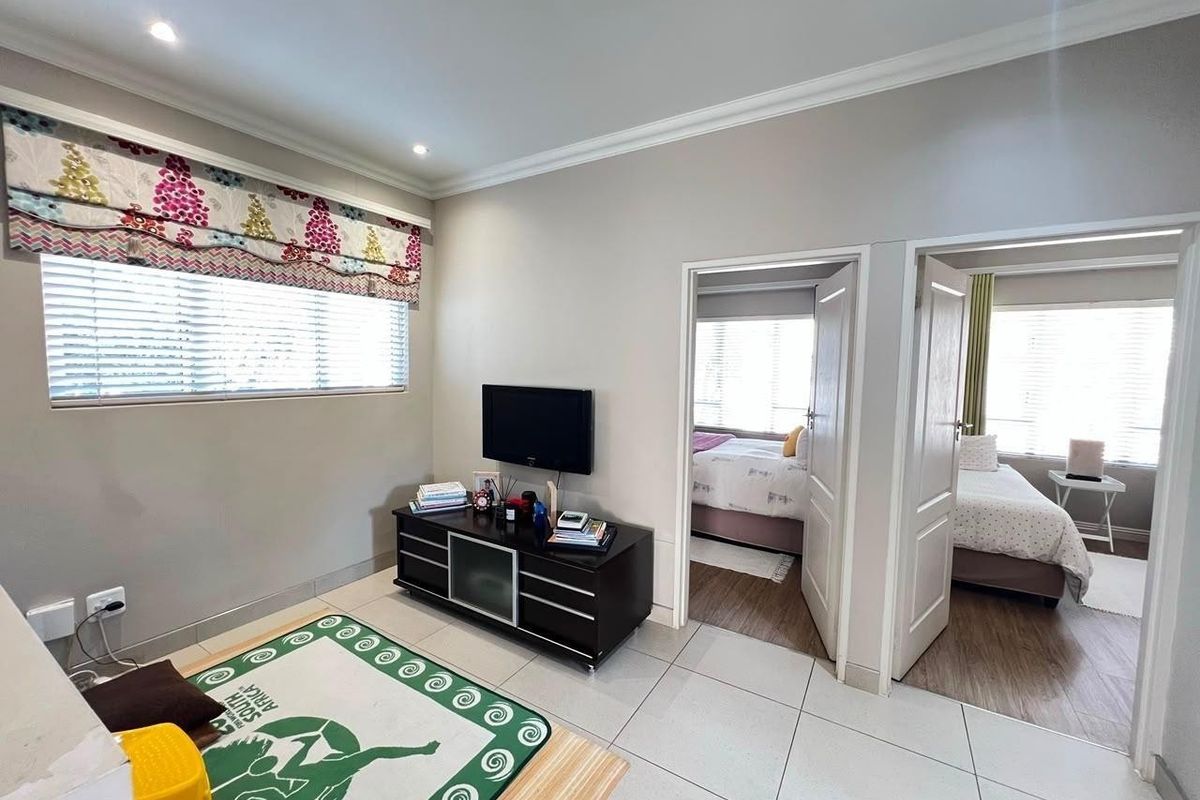 A bright living area featuring neutral tiled flooring, recessed downlighting, and access to two adjacent bedrooms through standard interior doors.
