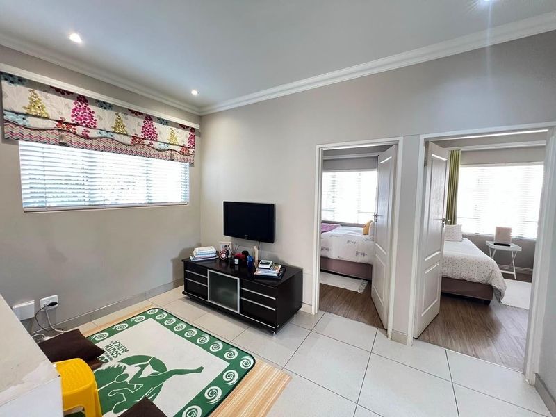 A bright living area featuring neutral tiled flooring, recessed downlighting, and access to two adjacent bedrooms through standard interior doors.