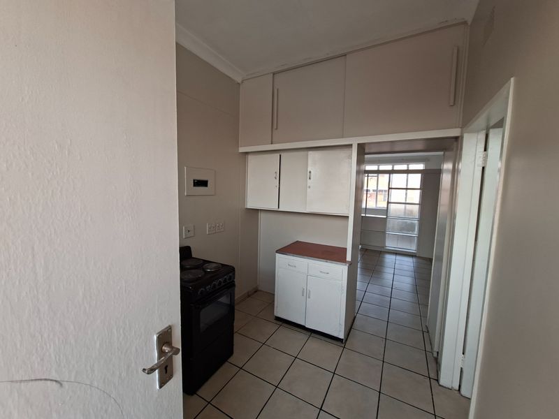 A compact, empty kitchen featuring tiled flooring, built-in cabinetry with overhead storage, and a freestanding stove, leading into an adjacent living area.