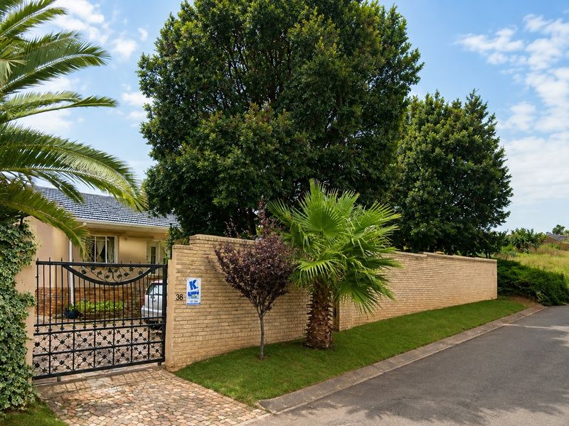 Front view of the property featuring a secure wrought iron gate, brick boundary wall, paved driveway, and established garden landscaping.