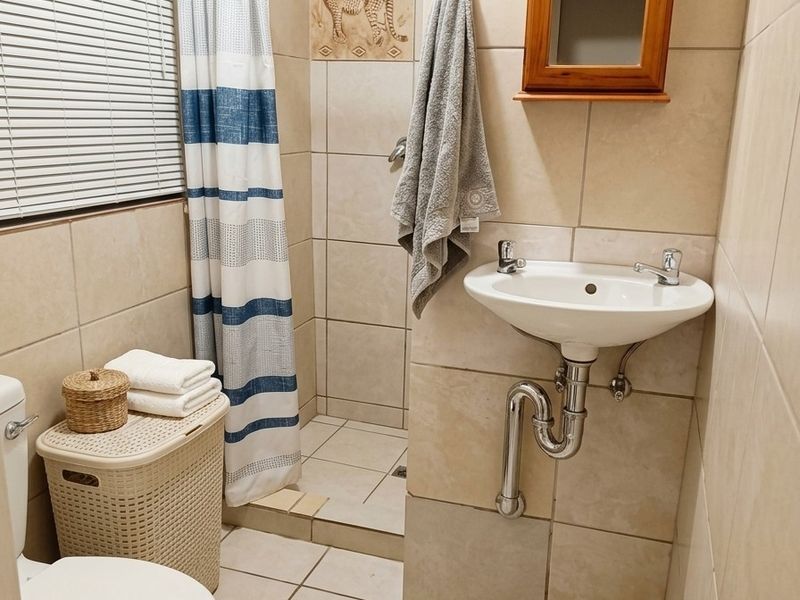 A compact bathroom featuring a wall-mounted basin, a shower cubicle with a curtain, and neutral floor-to-ceiling tiling.