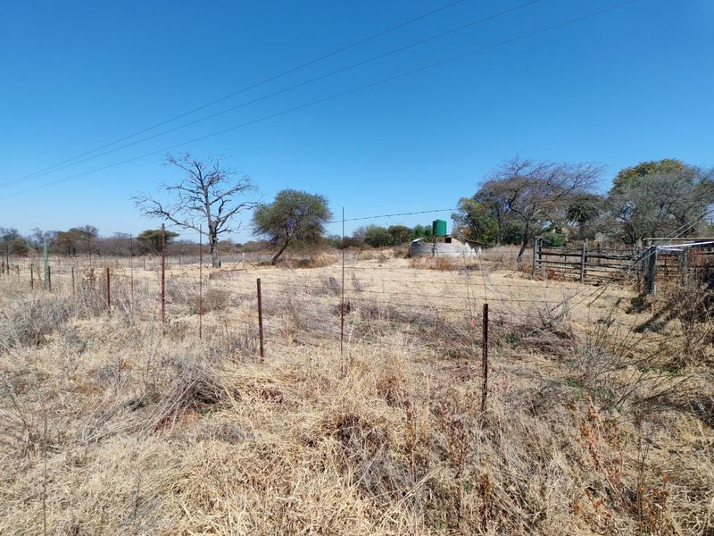 A view of a vacant residential plot featuring dry grass, perimeter barbed wire fencing, a water storage tank, and existing metal gate infrastructure.