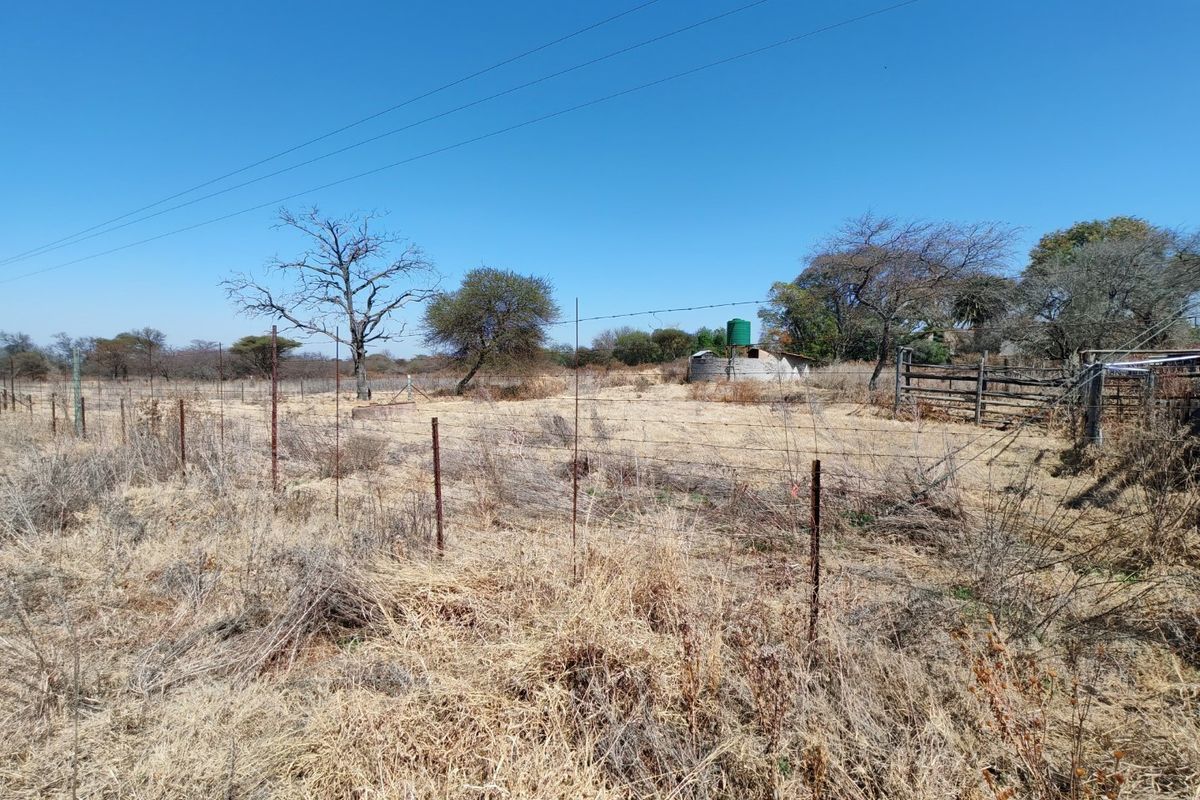 A view of a vacant residential plot featuring dry grass, perimeter barbed wire fencing, a water storage tank, and existing metal gate infrastructure.