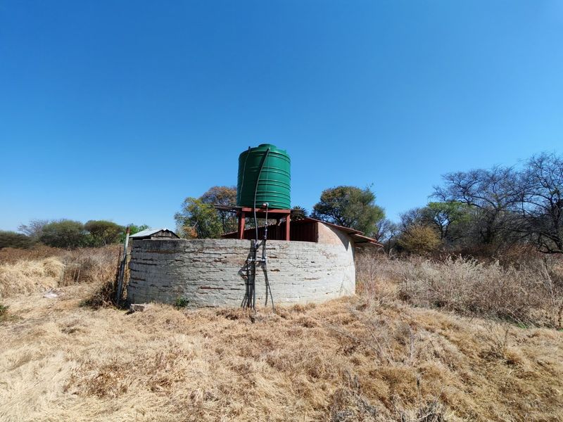 A circular brick utility structure featuring an elevated green water storage tank and associated plumbing infrastructure on a vacant residential plot.