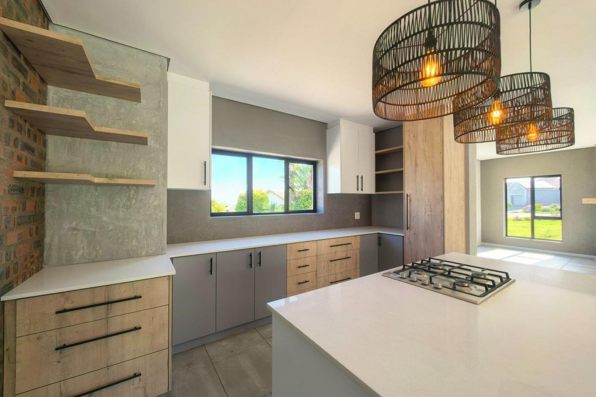 Modern kitchen featuring a central island with gas hob, sleek two-tone cabinetry, stone countertops, and an exposed brick feature wall with floating shelving.