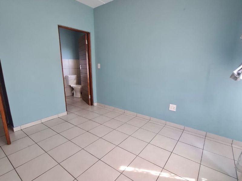 An empty bedroom featuring light blue walls and neutral ceramic floor tiles, with an adjoining en-suite bathroom visible through the doorway.