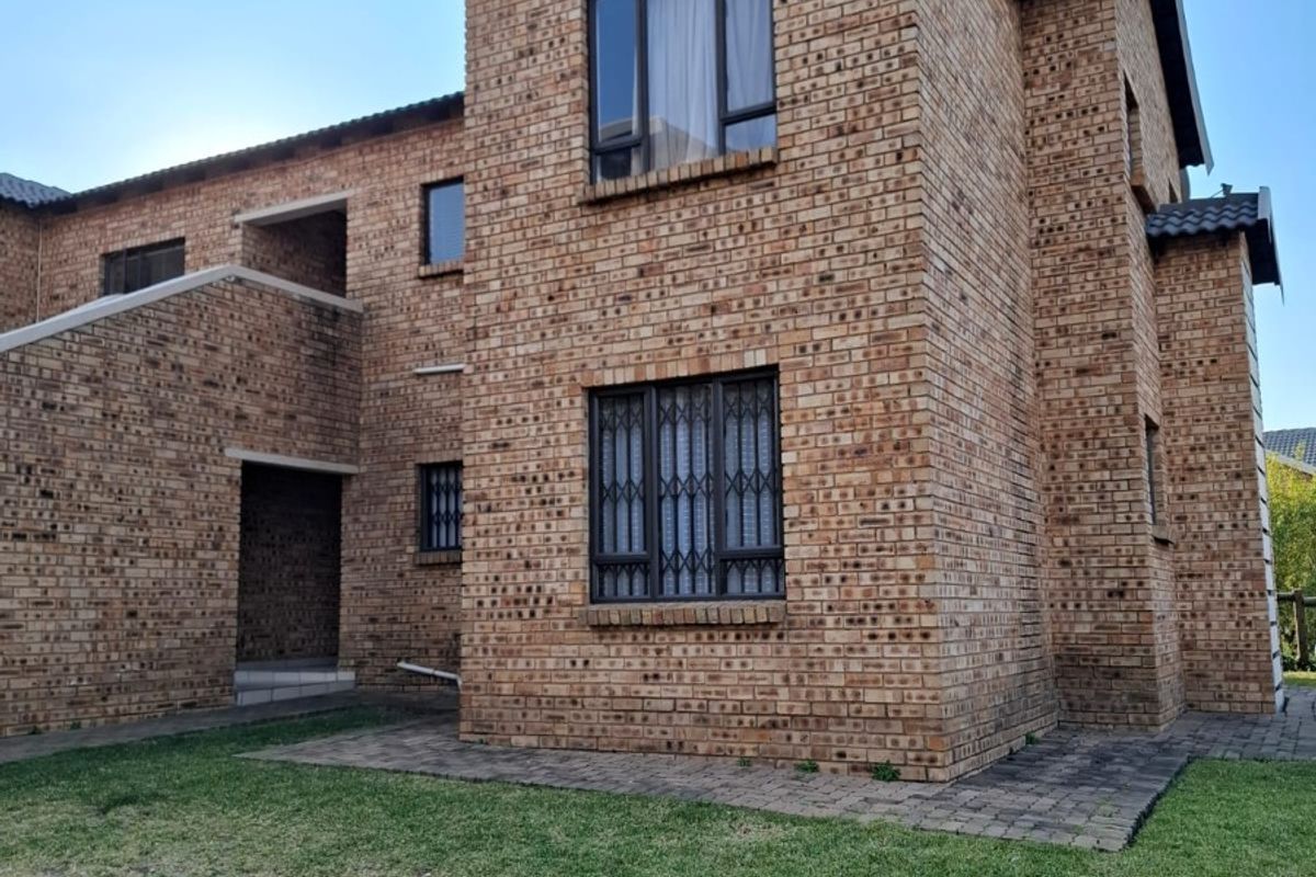 A two-story face brick apartment building exterior featuring aluminium window frames, security bars, and a surrounding lawn area with paved pathways.