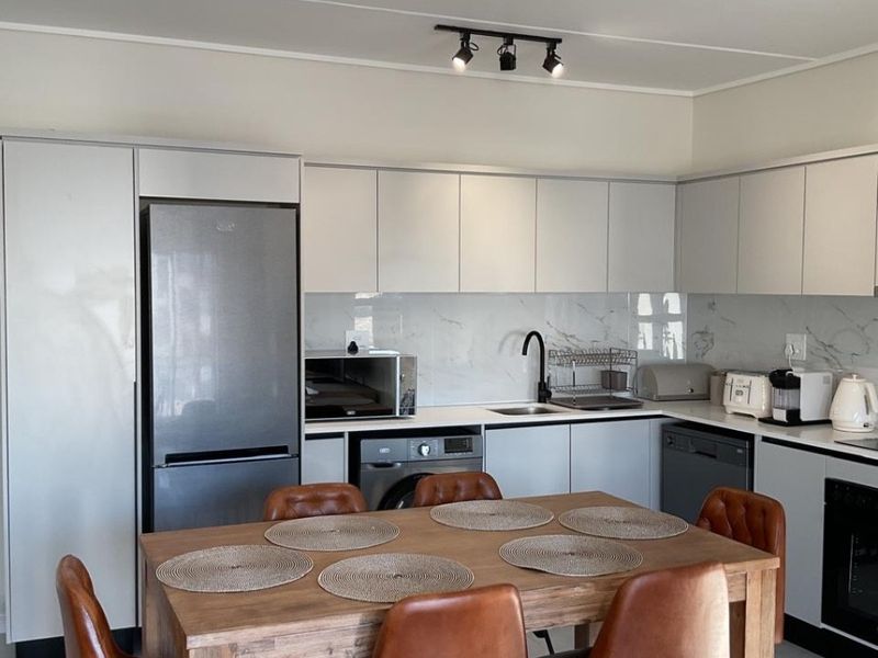 Modern open-plan kitchen and dining area featuring sleek white cabinetry, marble-look splashbacks, contemporary track lighting, and durable tiled flooring.