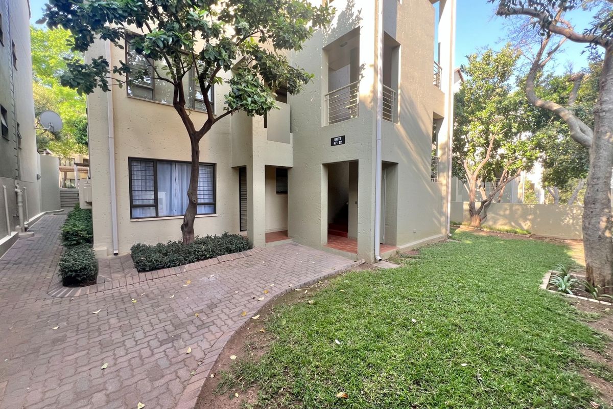 A view of the apartment complex exterior featuring a paved walkway, landscaped garden area, and access to upper-level units via external staircases.