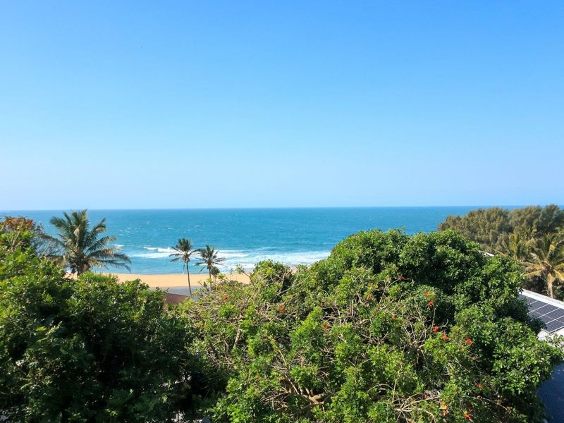 Elevated coastal view overlooking the ocean, sandy beach, and surrounding lush greenery, featuring visible solar panels on the adjacent roof structure.