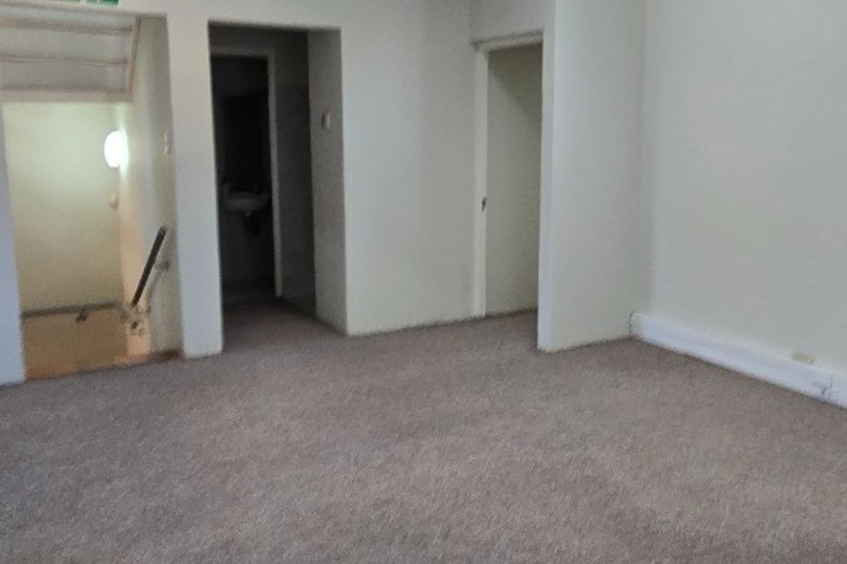 An empty, carpeted living area featuring a wall-mounted air conditioning unit, fluorescent lighting, and access to a staircase and adjacent rooms.