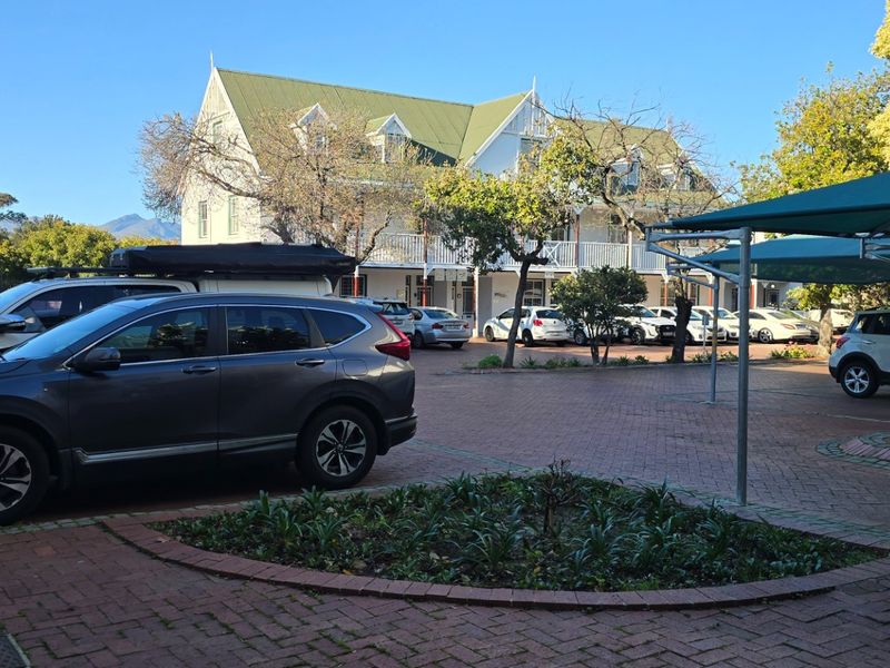 A paved parking area featuring a covered carport structure and established garden beds in front of a multi-story residential building.