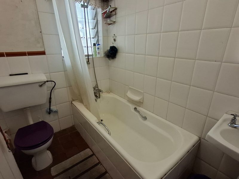 A dated bathroom featuring a standard bathtub, pedestal sink, and toilet with white wall tiling and a louvered window.