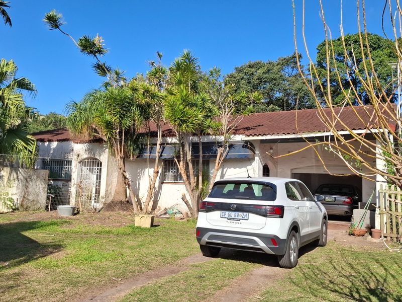 Front elevation of a single-story house featuring a carport, security bars on windows, and a tiled roof, set within a garden area.