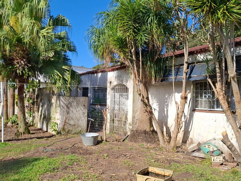 Rear exterior view of the house showing weathered masonry, security bars on windows, and a tiled roof structure set within a garden area.