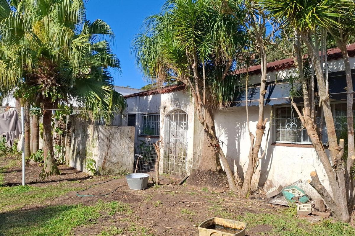 Rear exterior view of the house showing weathered masonry, security bars on windows, and a tiled roof structure set within a garden area.