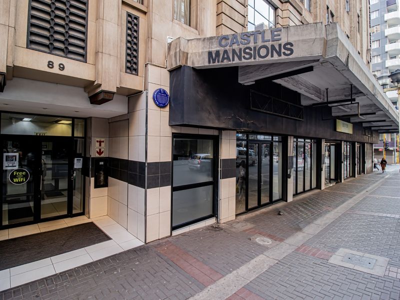 Street-level view of the Castle Mansions commercial building entrance, featuring a prominent canopy, glass storefronts, and tiled exterior facade.