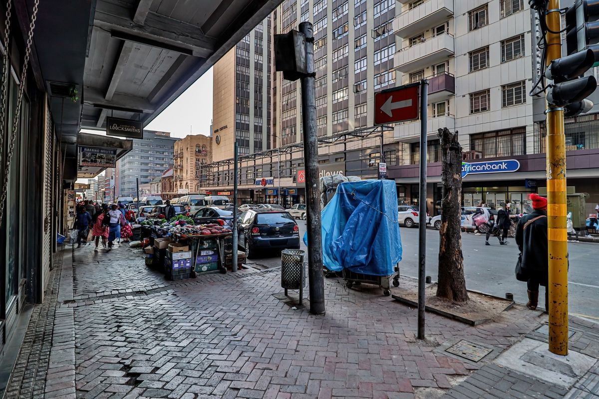 A street-level view of a busy urban commercial area featuring paved sidewalks, traffic infrastructure, and surrounding multi-story commercial buildings.